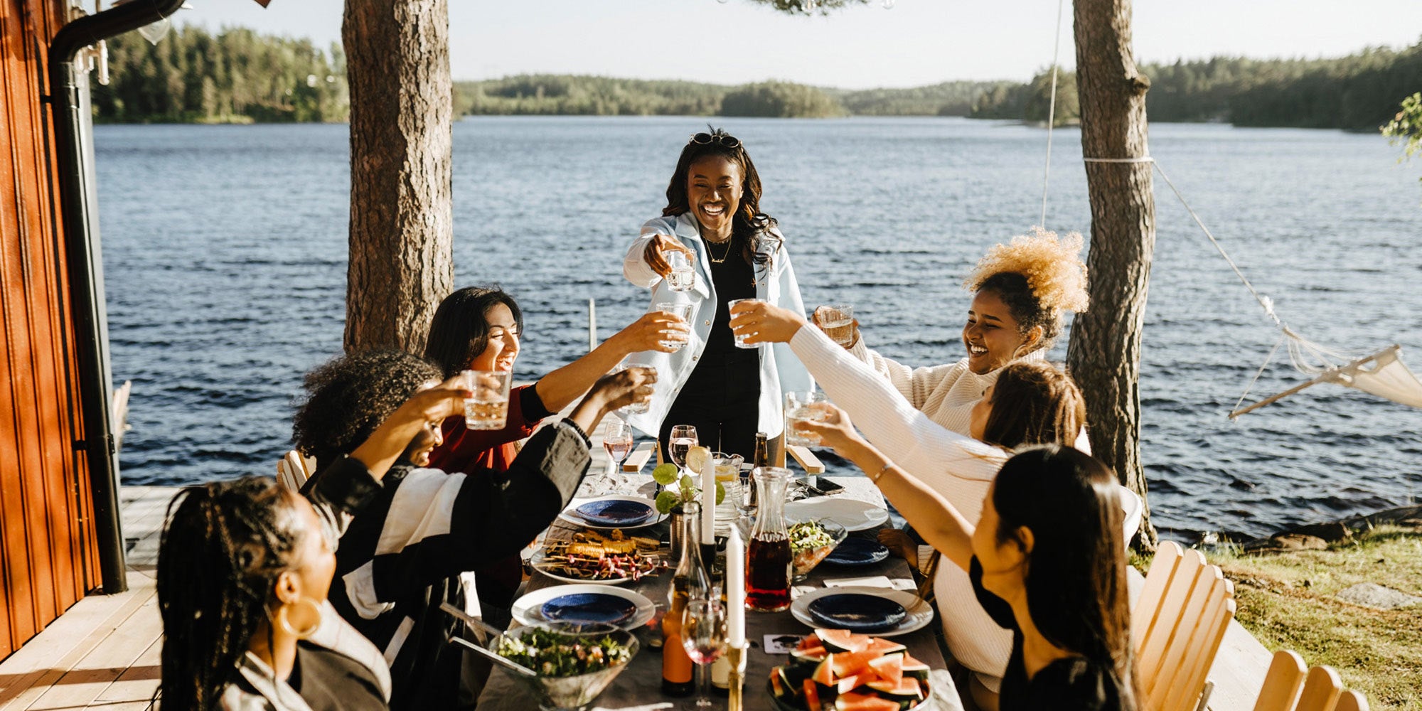 Group of friends sit around a table by the water and raise their glasses in a toast 