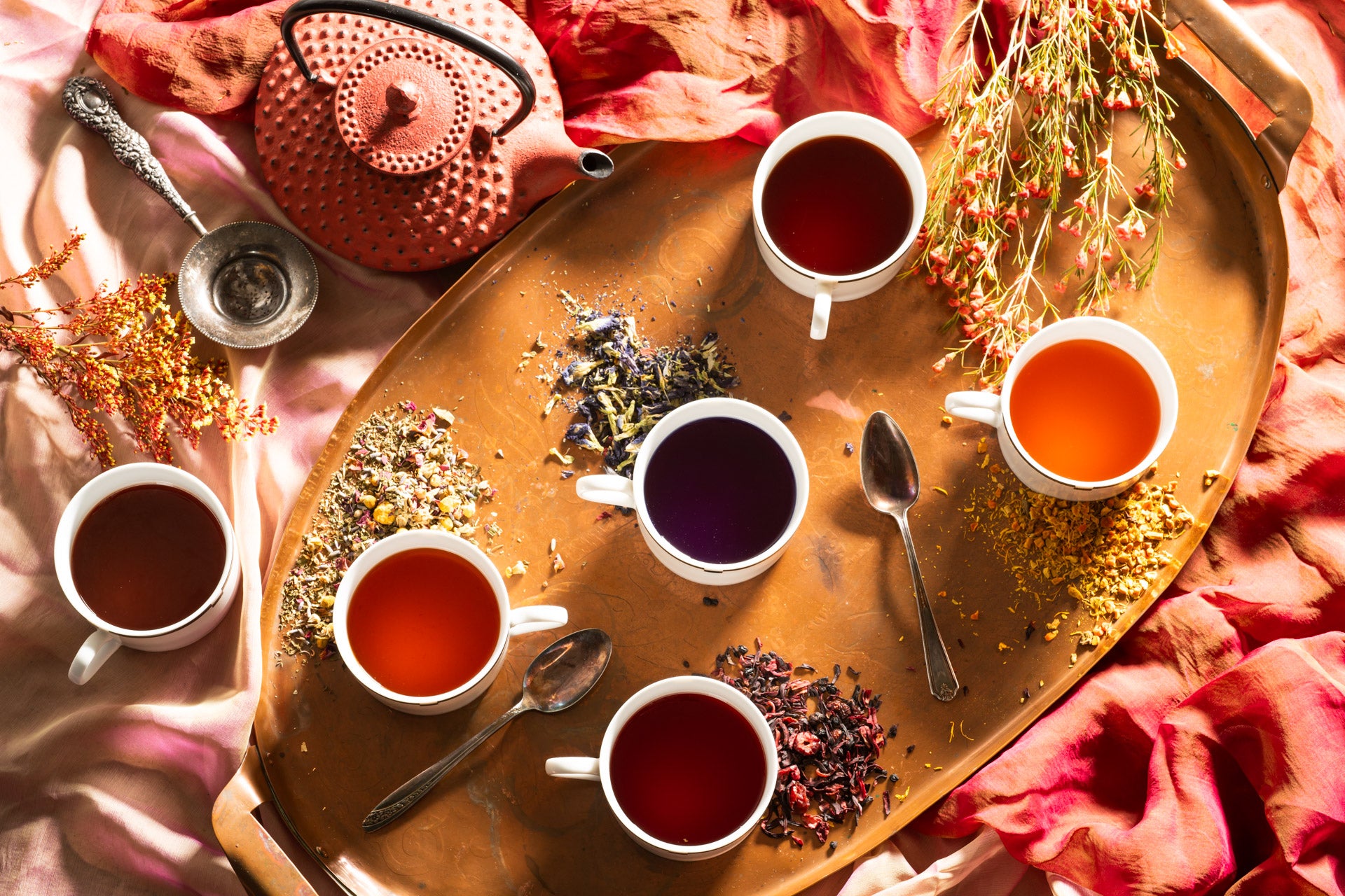 cups of tea on a bronze tray with spoons, a red tea kettle, and loose leaf tea around 